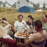 Autour d'une table, un groupe d'adulte déjeune. La photo a du grain et parait un peu vieillie. Il y a du vent et peu de soleil. Derrière, des herbes folles, une tente bleue, puis des immeubles. Les gens sourient en regardant l'objectif.