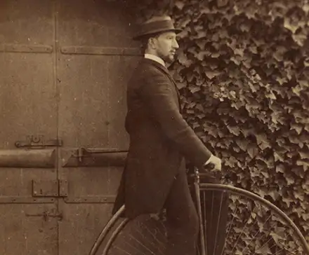 Une photographie en noir et blanc d'un homme en costume. Il se tient de profil sur un  grand bi, une bicyclette qui possède une roue avant d'un très grand diamètre et une roue arrière beaucoup plus petite. Il pose devant une porte et un mur recouvert de lierre.