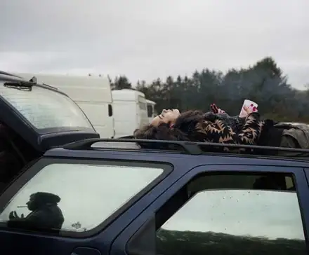 une femme est allongée sur le dos, sur le toit d'une voiture entourée d'autres véhicules. Elle tient dans sa main un gobelet et une bouteille.