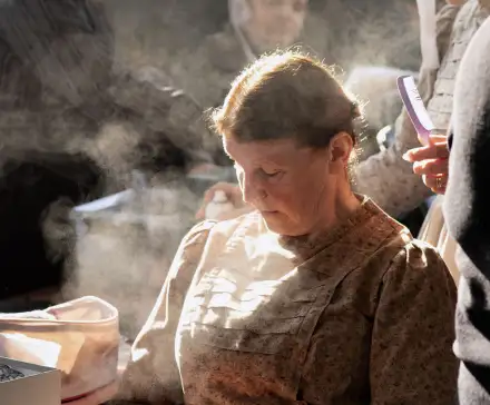 Photographie en couleur. Au premier plan, une femme en costume breton est assise, entourée d'autres femmes. Elle est auréolée d'un rayon de lumière et entourée de vapeurs de laques. La femme derrière elle tient dans sa main un peigne.