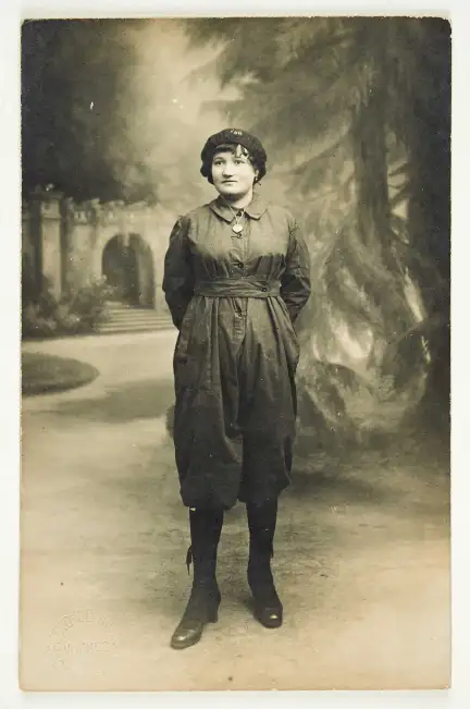 Une jeune femme en tenue de travail de la poudrerie pose devant une toile peinte. Elle est vêtue d'un béret, et d'une combinaison dont les jambes s'arrêtent aux genoux.